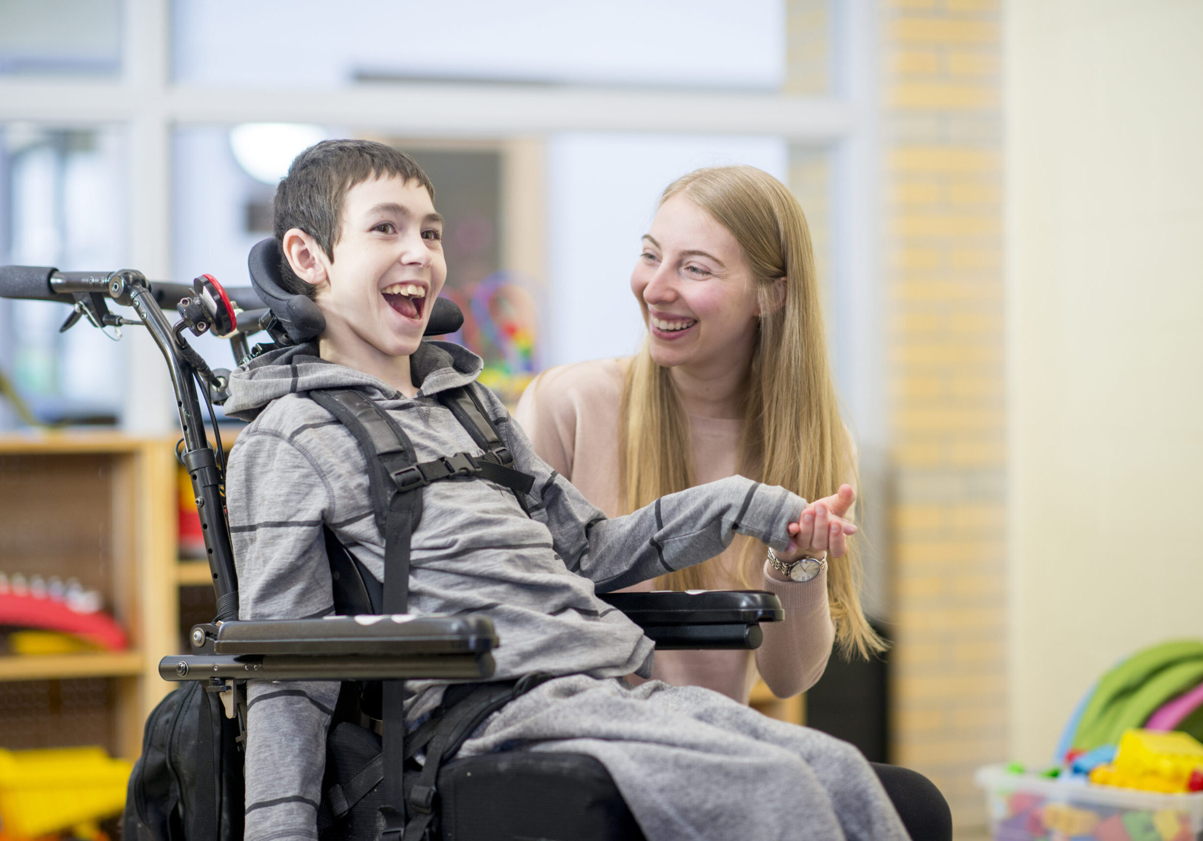 A caregiver is playing with a little boy that has a mental and physical disability. He is happily sitting in his wheelchair.